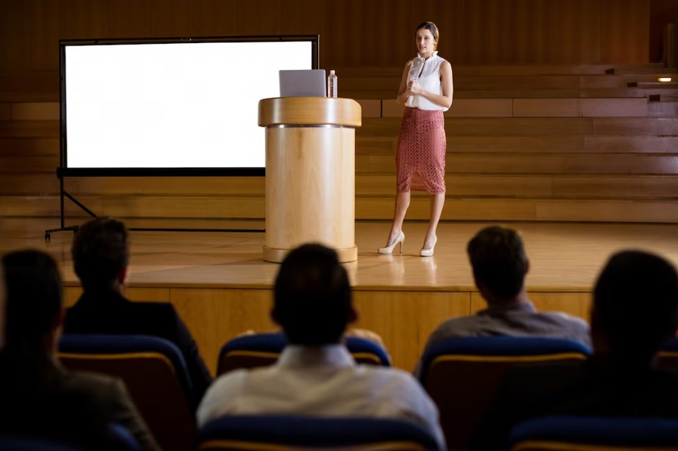 a businesswoman giving a presentation on stage in front of a seated audience