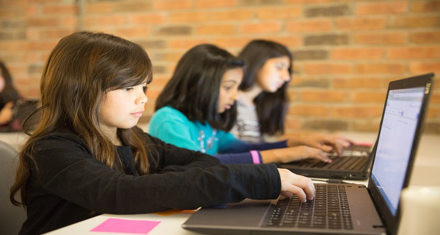 young girls are sitting at tables and working with laptops